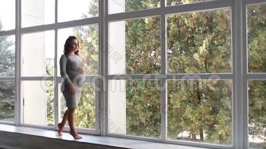 美丽的孕妇走在房子里，靠近一扇大窗户。 她穿着孕妇装. 美丽美丽